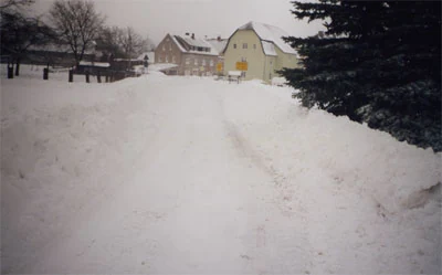 Eine verschneite Straße führt zwischen hohen Schneerändern auf Häuser zu.